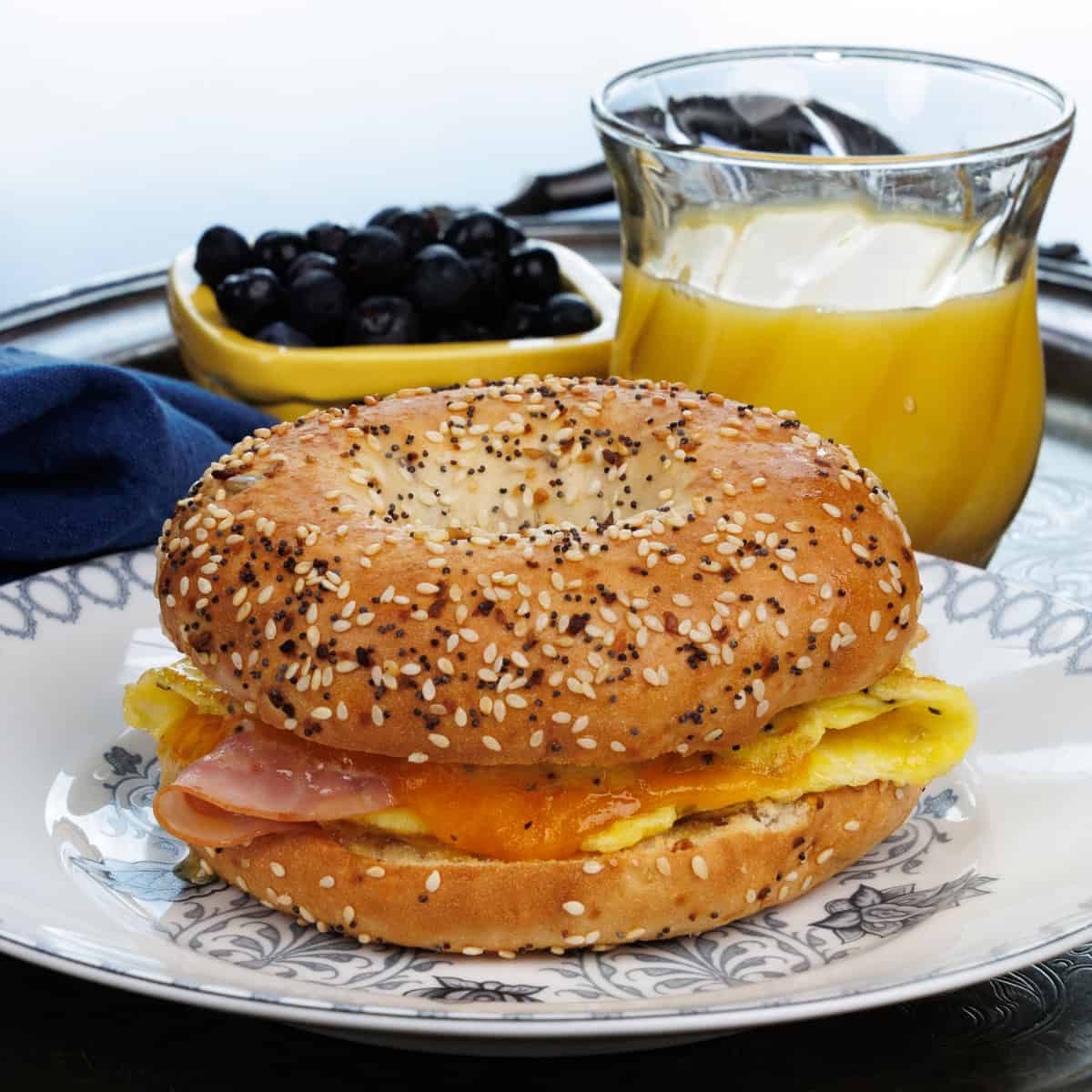 a ham and cheese breakfast bagel on a white plate next to a bowl of blueberries.