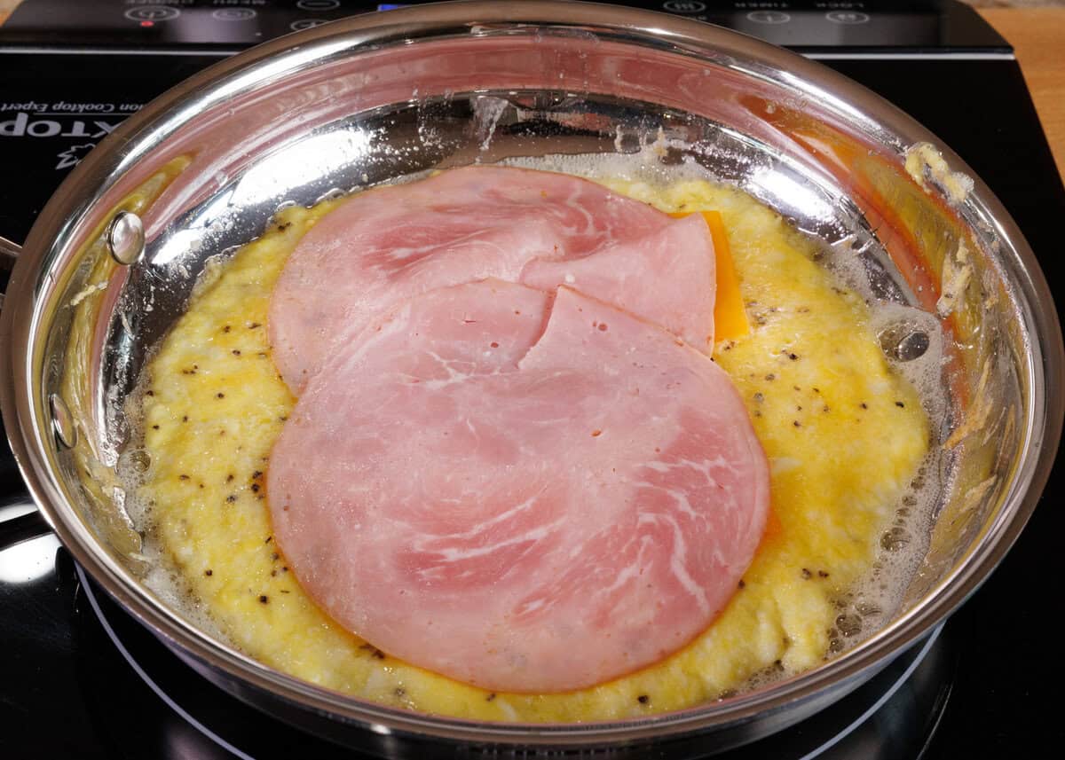 a slice of ham and a slice of cheese on top of cooked eggs in a skillet.