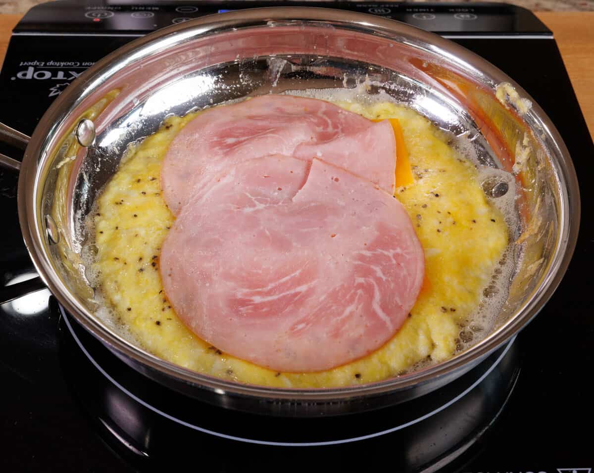 a slice of ham and a slice of cheese on top of cooked eggs in a skillet.