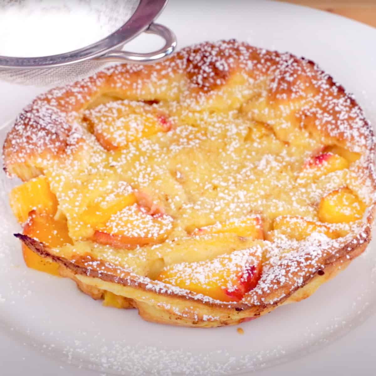 a single serve peach dutch baby topped with powdered sugar on a white plate.