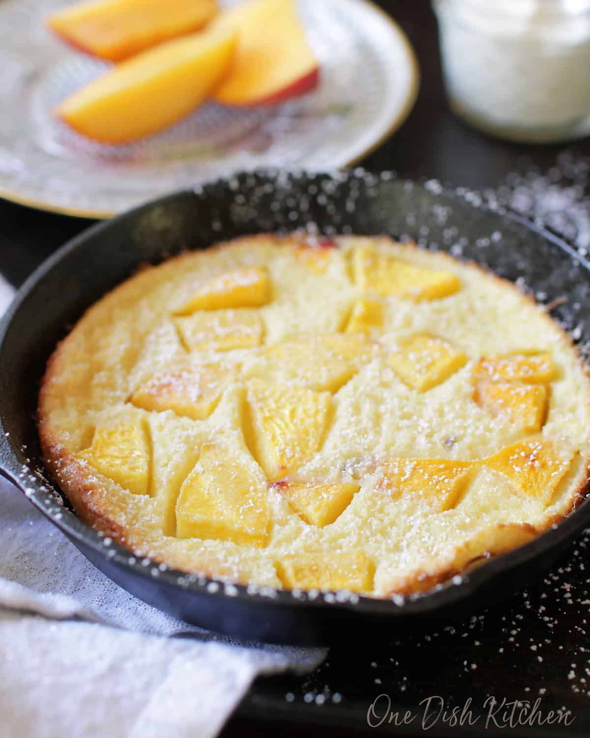 A puffed pancake in a small cast iron skillet topped with chopped peaches and dusted with powdered sugar with a small jar of whipped cream and a plate of sliced peaches in the background.