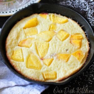 A puffed pancake in a small cast iron skillet topped with chopped peaches and dusted with powdered sugar with a small jar of whipped cream and a plate of sliced peaches in the background.