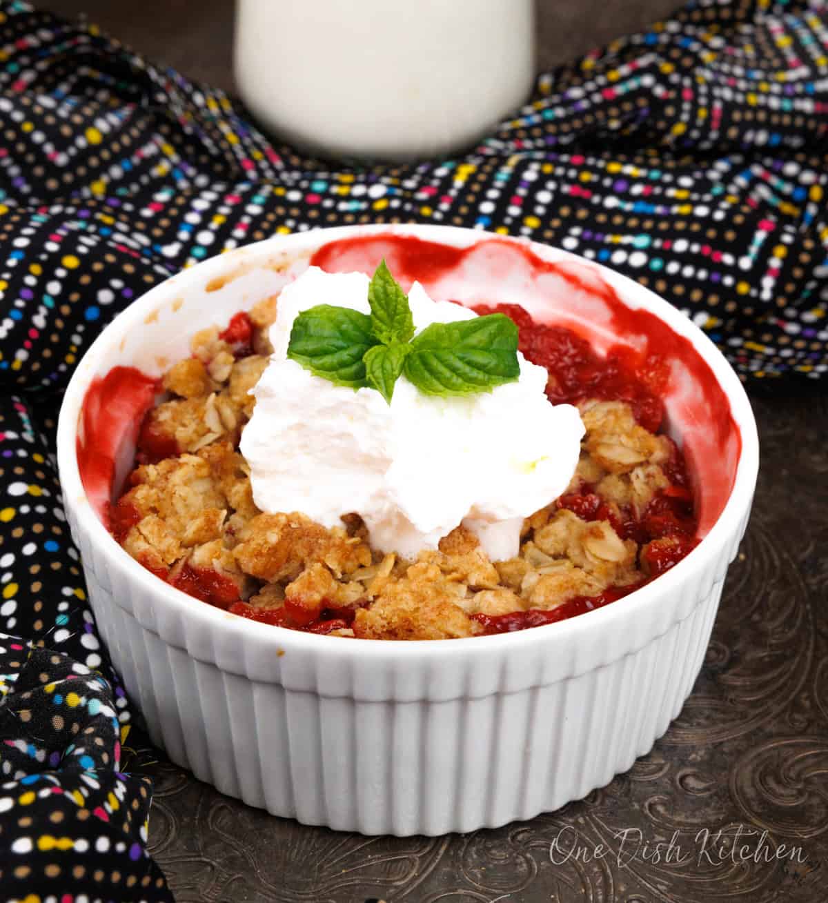 a single serving strawberry crisp with juicy strawberries under an oat topping in a ramekin topped with whipped cream.