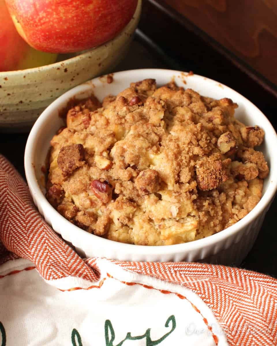 a bakery style apple muffin topped with a pecan streusel topping next to a bowl of apples and an orange napkin.