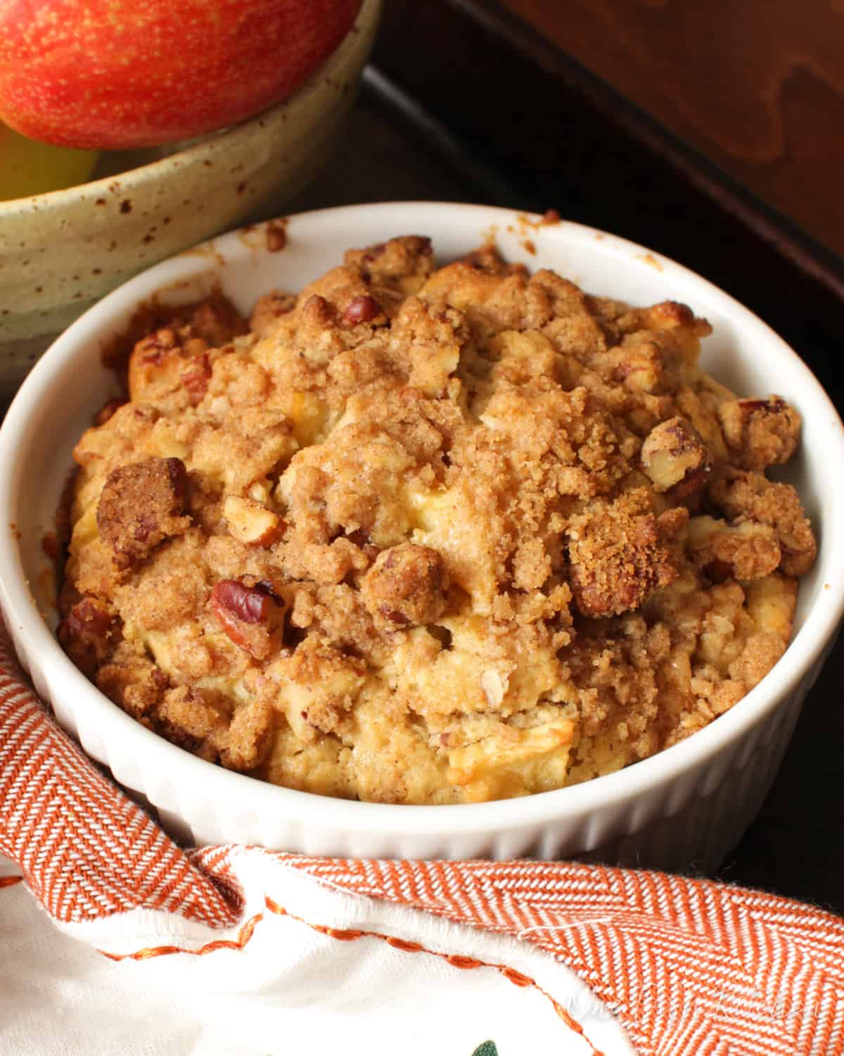 a bakery style apple muffin topped with a pecan streusel topping next to a bowl of apples and an orange napkin.