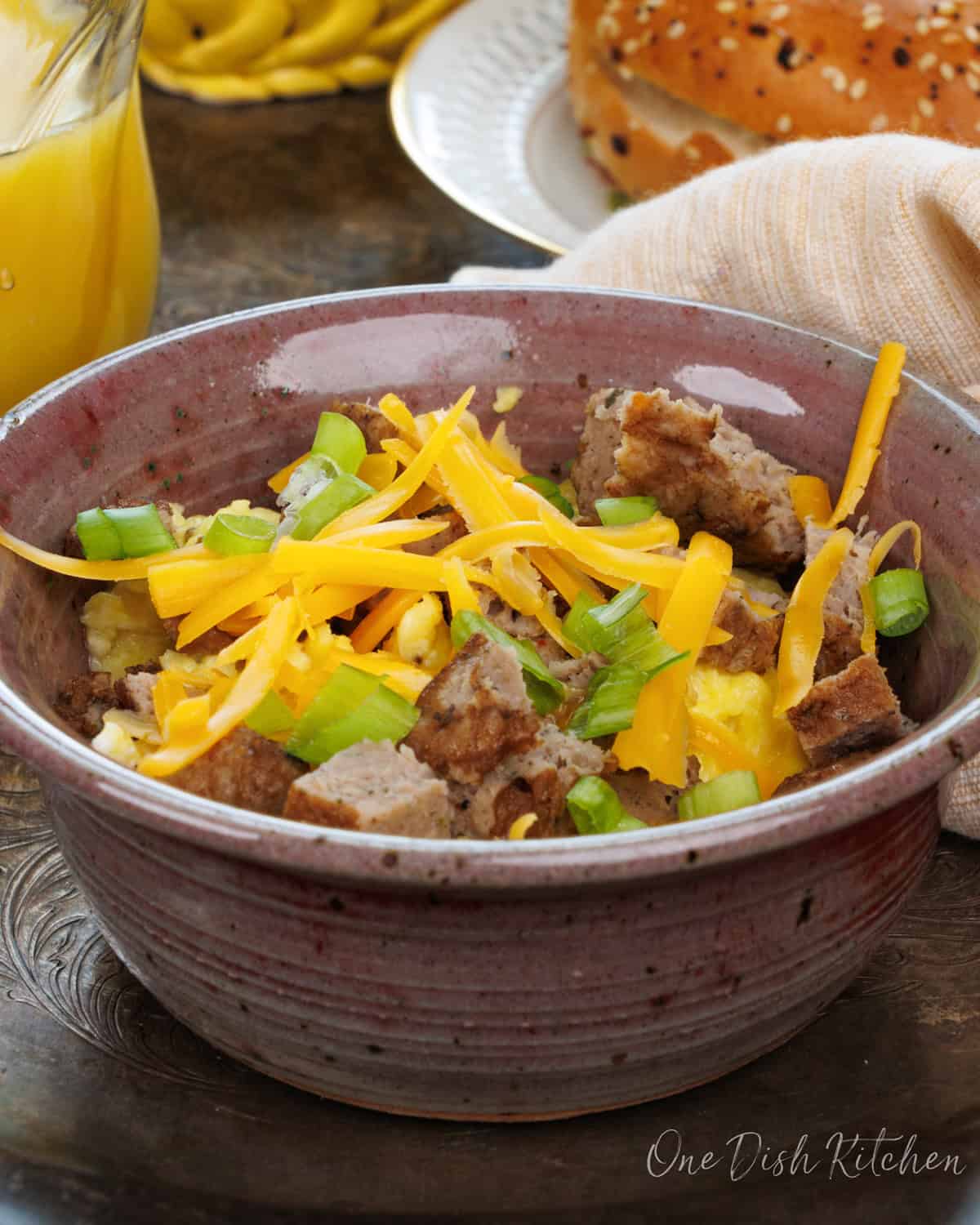 a breakfast bowl filled with eggs, sausage, cheese and topped with green onions on a silver tray next to a bagel.