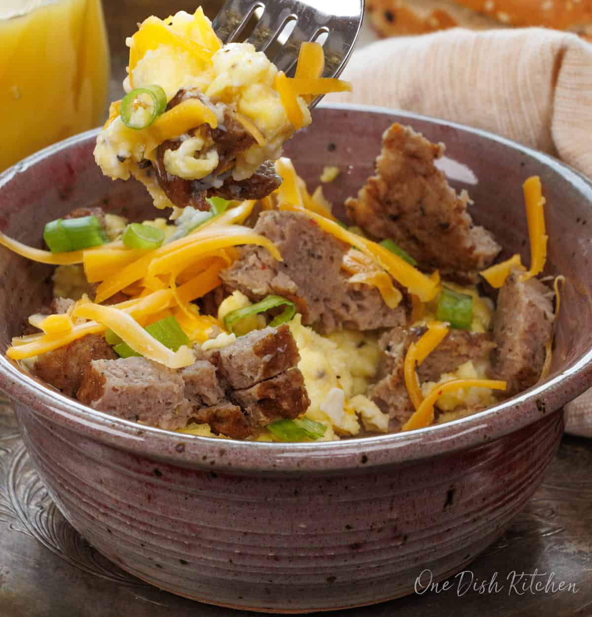 a fork filled with scrambled eggs and sausage over a bowl on a silver tray.