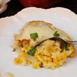 a scoop of corn casserole on a white plate with a spoon on the plate next to a casserole dish filled with the remaining corn casserole.