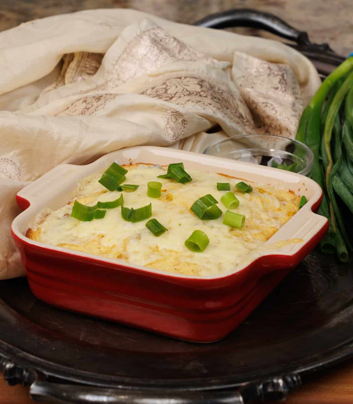 a small corn casserole topped with chopped green onions on a silver tray next to a cream colored napkin