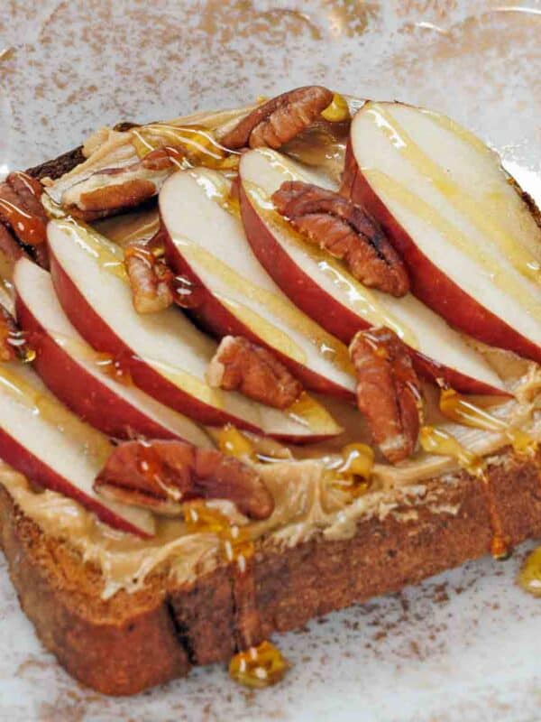 peanut butter on a slice of toast topped with apple slices and a drizzle of honey on a white plate next to a blue jar of honey.