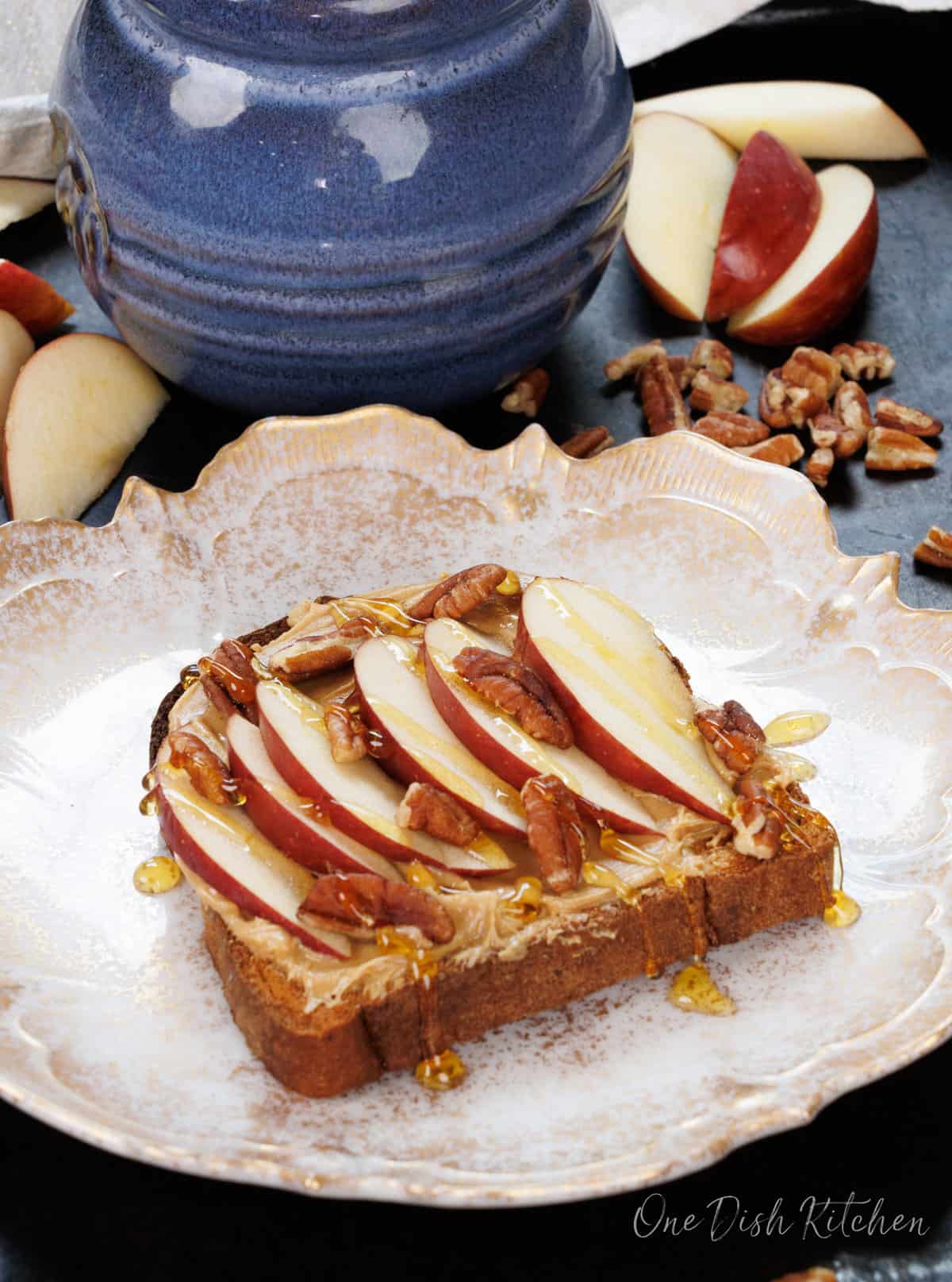 peanut butter on a slice of toast topped with apple slices and a drizzle of honey on a white plate next to a blue jar of honey