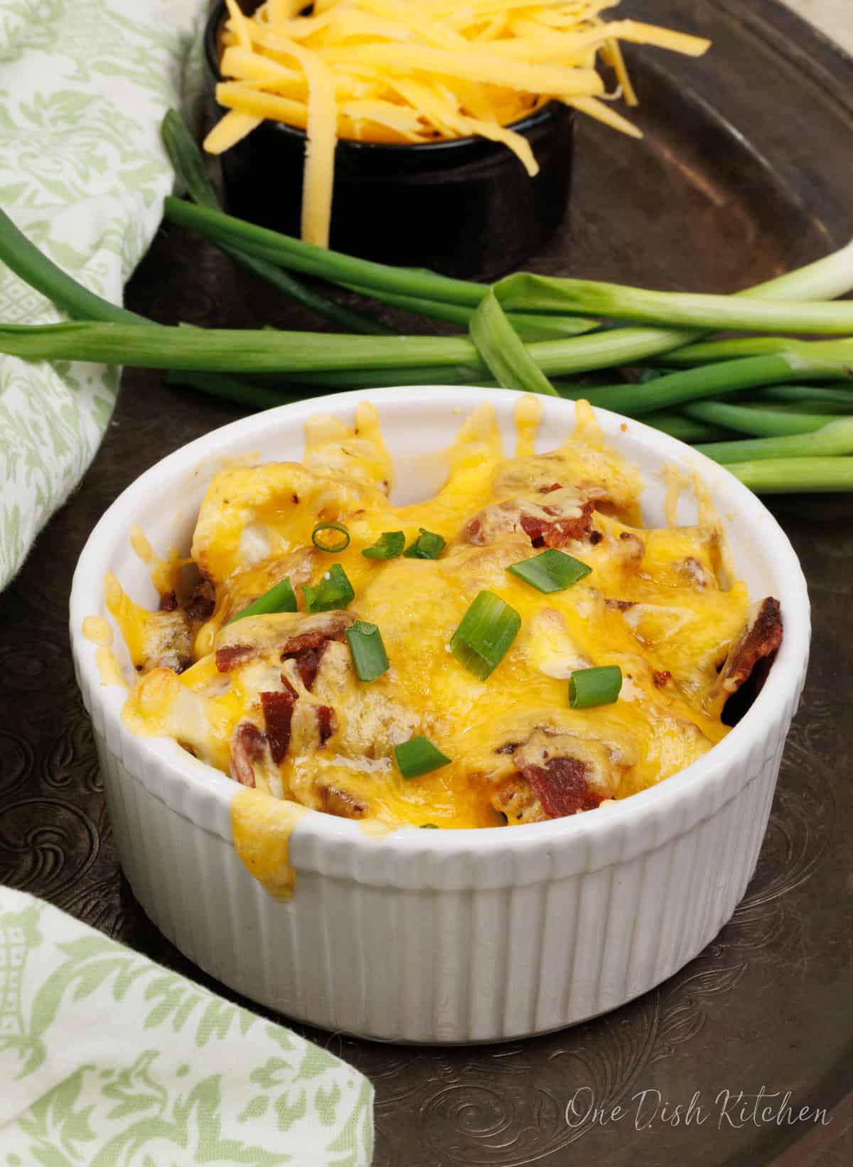 a mini cauliflower casserole topped with green onions next to a bowl of shredded cheddar cheese and a green napkin