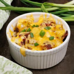 a mini cauliflower casserole topped with green onions next to a bowl of shredded cheddar cheese and a green napkin