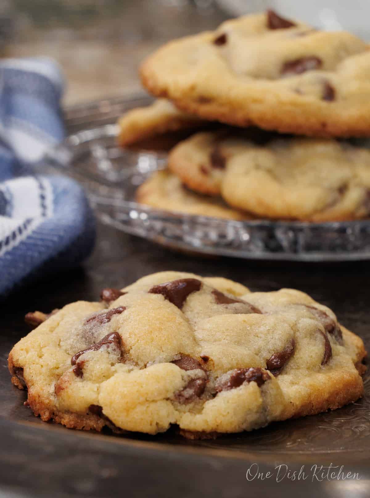 one chocolate chip cookie on a silver tray next to a plate of four chocolate chip cookies.