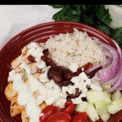 a red bowl filled with rice, chicken, kalamata olives, feta cheese, tomatoes, cucumbers and topped with tzatziki sauce