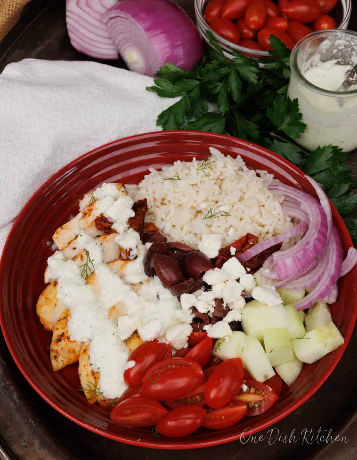 a red bowl filled with rice, chicken, kalamata olives, feta cheese, tomatoes, cucumbers and topped with tzatziki sauce