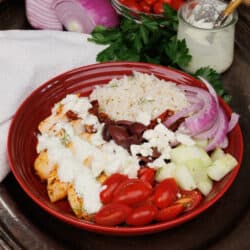 a greek chicken and rice bowl topped with tzatziki sauce on a silver tray next to a white napkin
