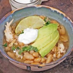 a bowl filled with slow cooker white bean and chicken chili topped with avocado slices and sour cream next to a bowl of sour cream.