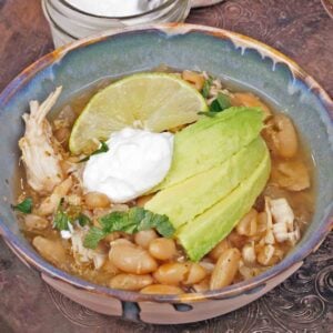 a bowl filled with slow cooker white bean and chicken chili topped with avocado slices and sour cream next to a bowl of sour cream.