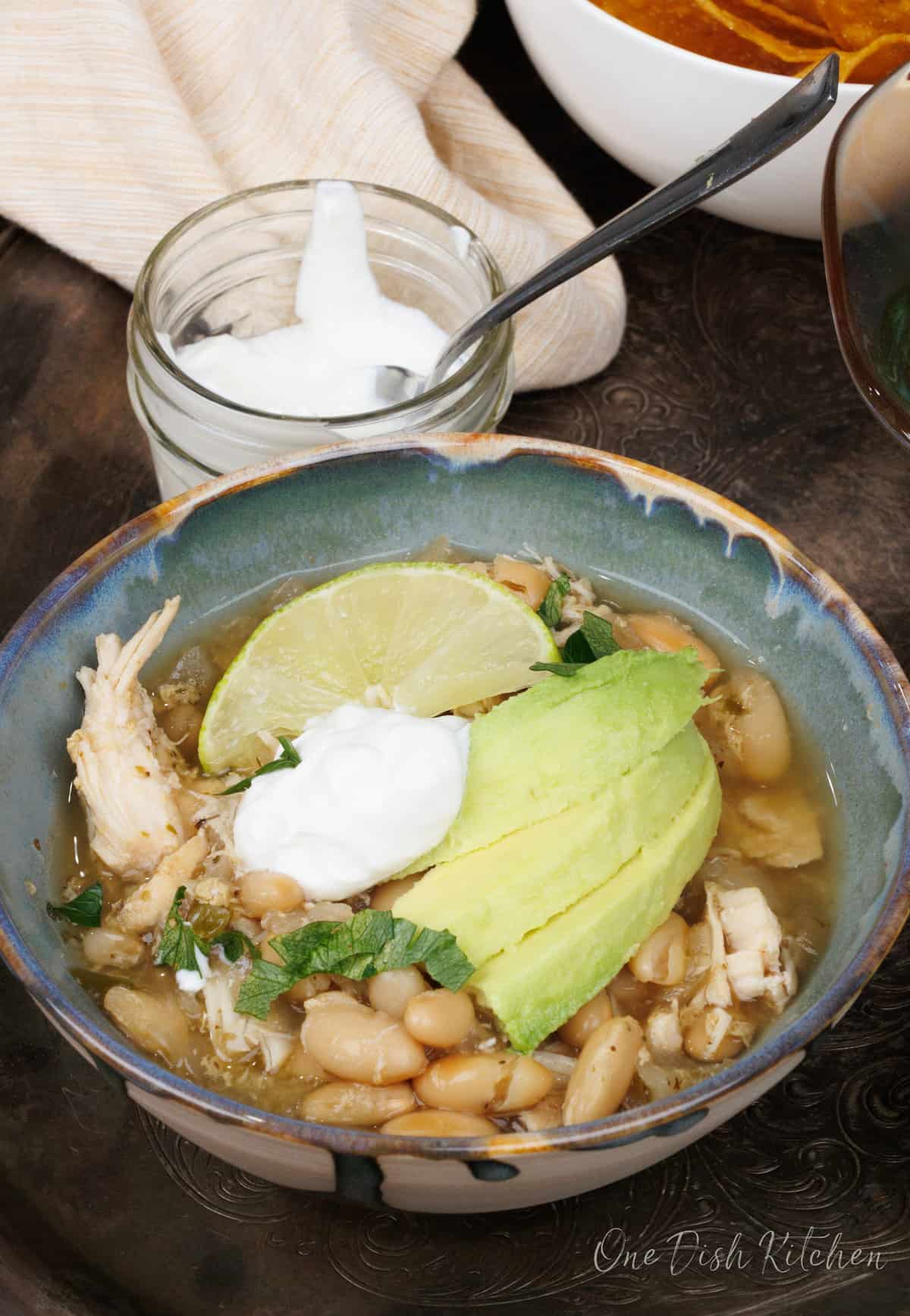 a bowl filled with white bean and chicken chili topped with avocado slices and sour cream next to a bowl of sour cream.