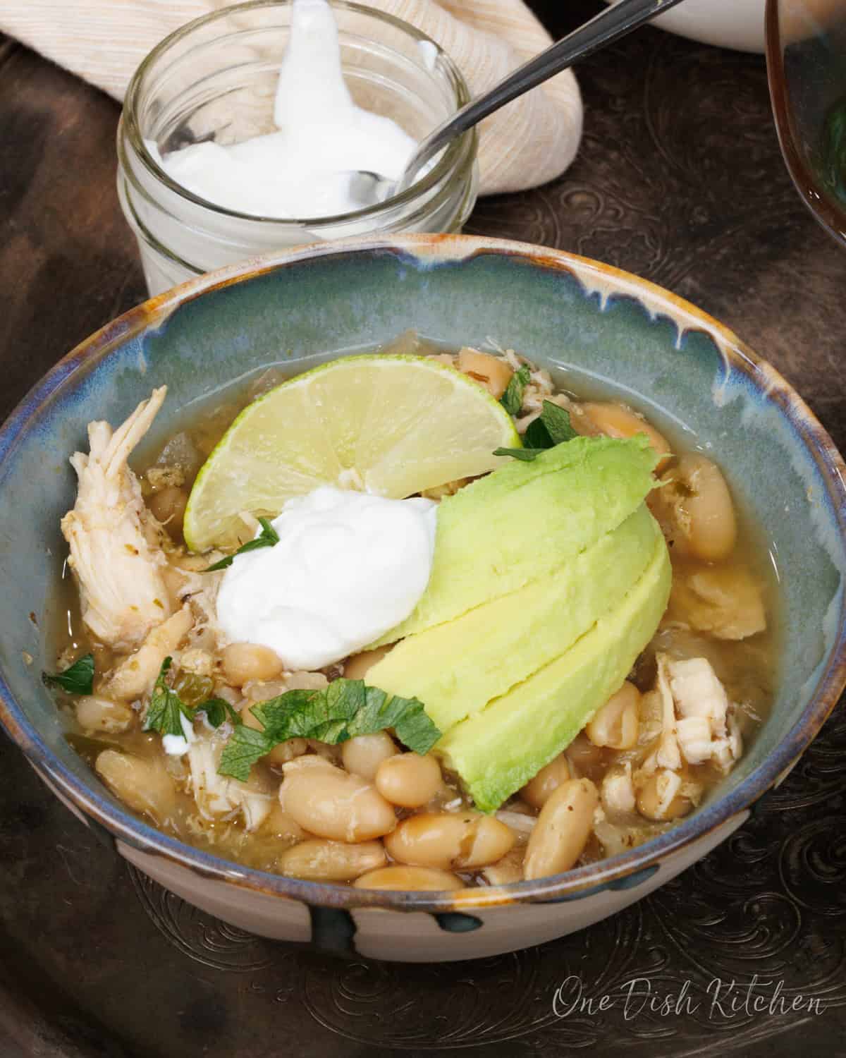 a bowl filled with white bean and chicken chili topped with avocado slices and sour cream next to a bowl of sour cream.