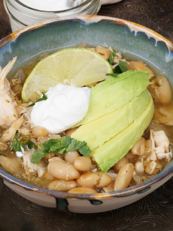 a bowl filled with slow cooker white bean and chicken chili topped with avocado slices and sour cream next to a bowl of sour cream