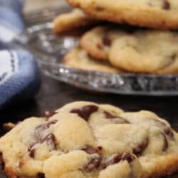 one chocolate chip cookie on a silver tray next to a plate of four chocolate chip cookies