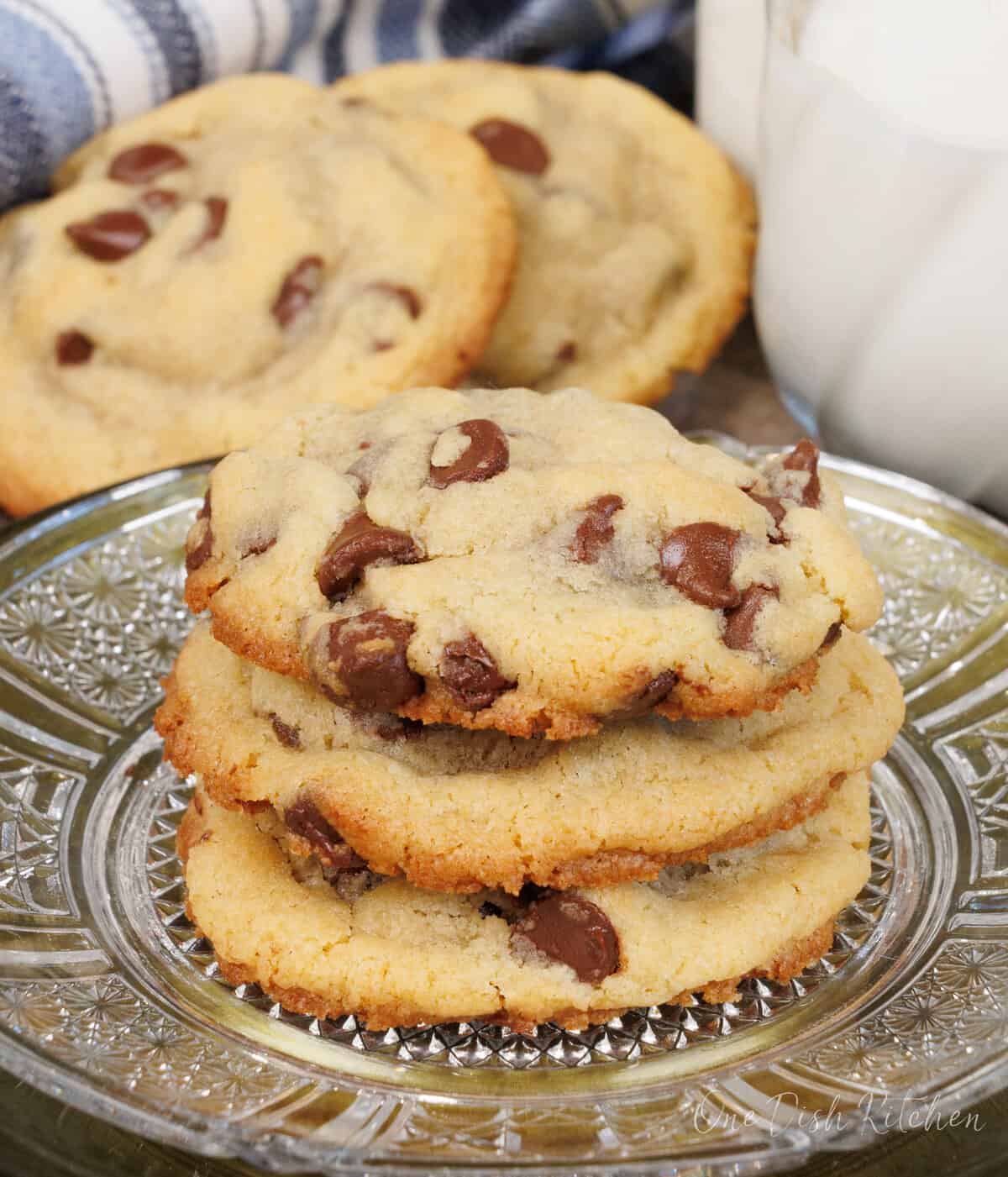 three chocolate chip cookies stacked on top of each other on a clear plate next to a glass of milk.