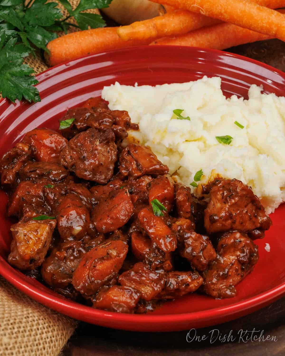 coq au vin in a red bowl next to mashed potatoes.