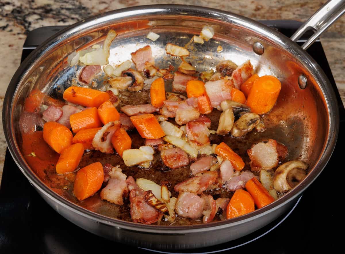 bacon, carrots, onions, and mushrooms cooking in a skillet on the stove