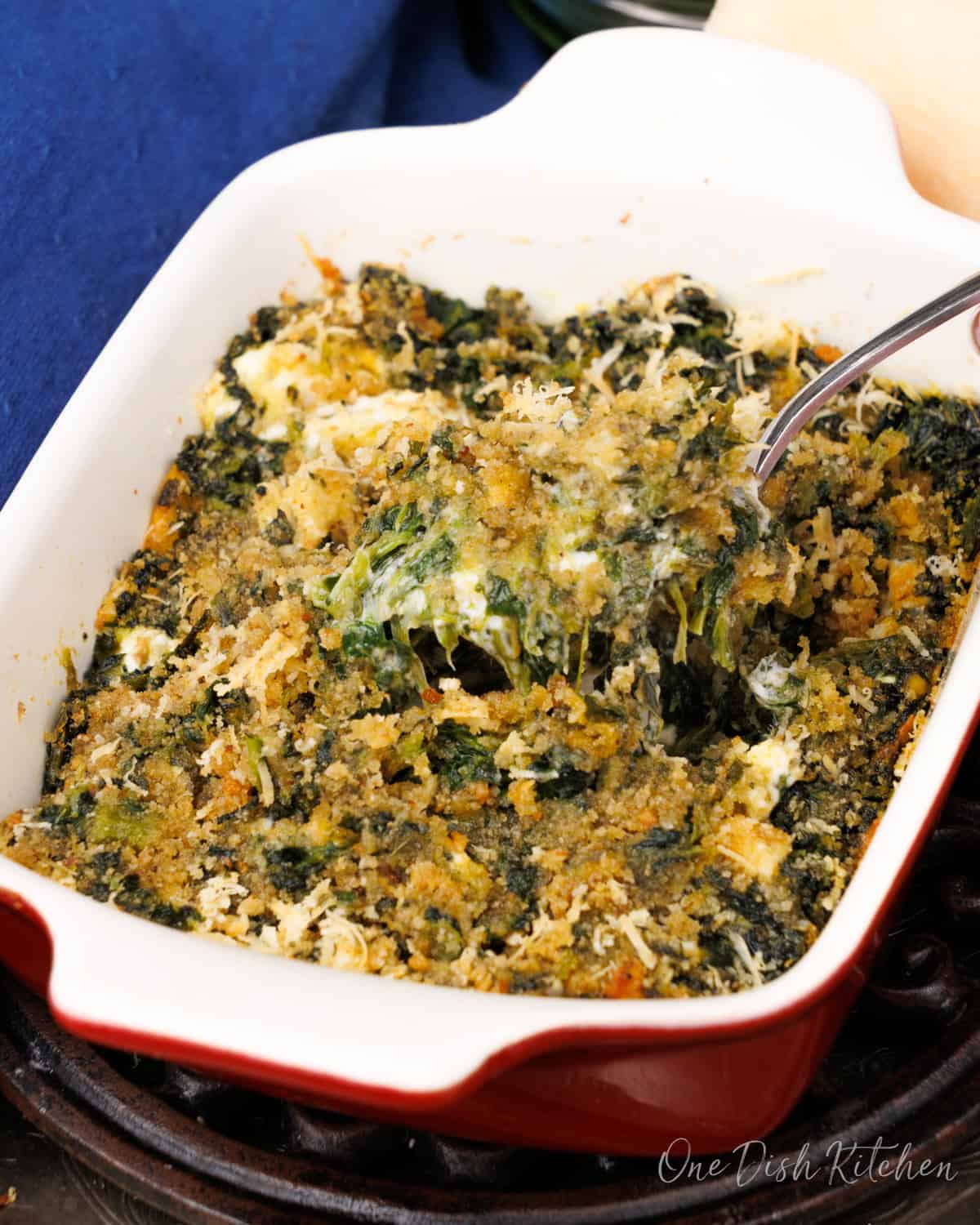 A small creamed spinach casserole in a red baking dish next to a blue napkin.