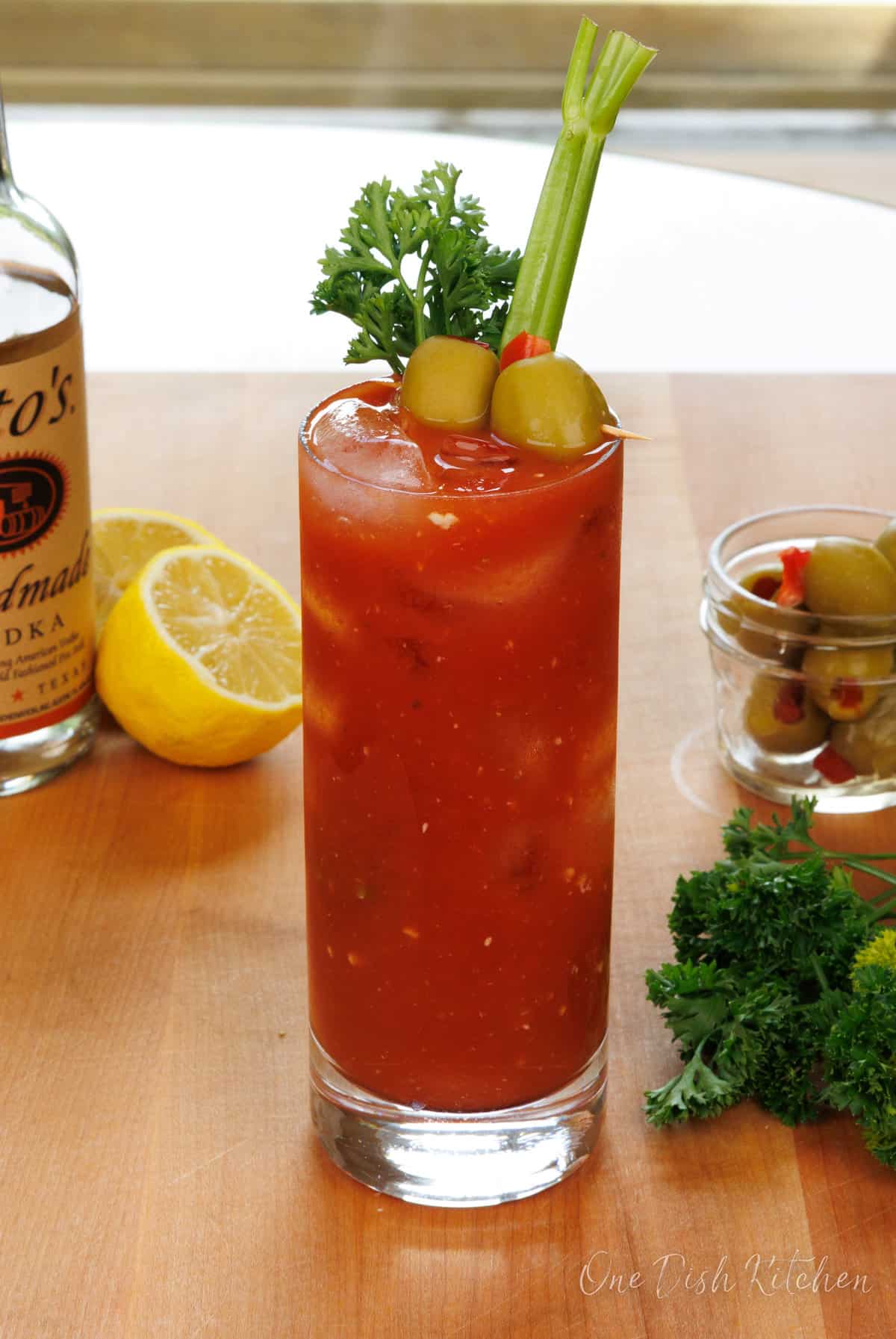 a bloody mary cocktail on a kitchen table next to a lemon wedge, olives, and celery