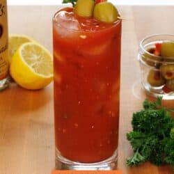 a bloody mary in a tall glass next to fresh parsley, olives, and lemon slices.