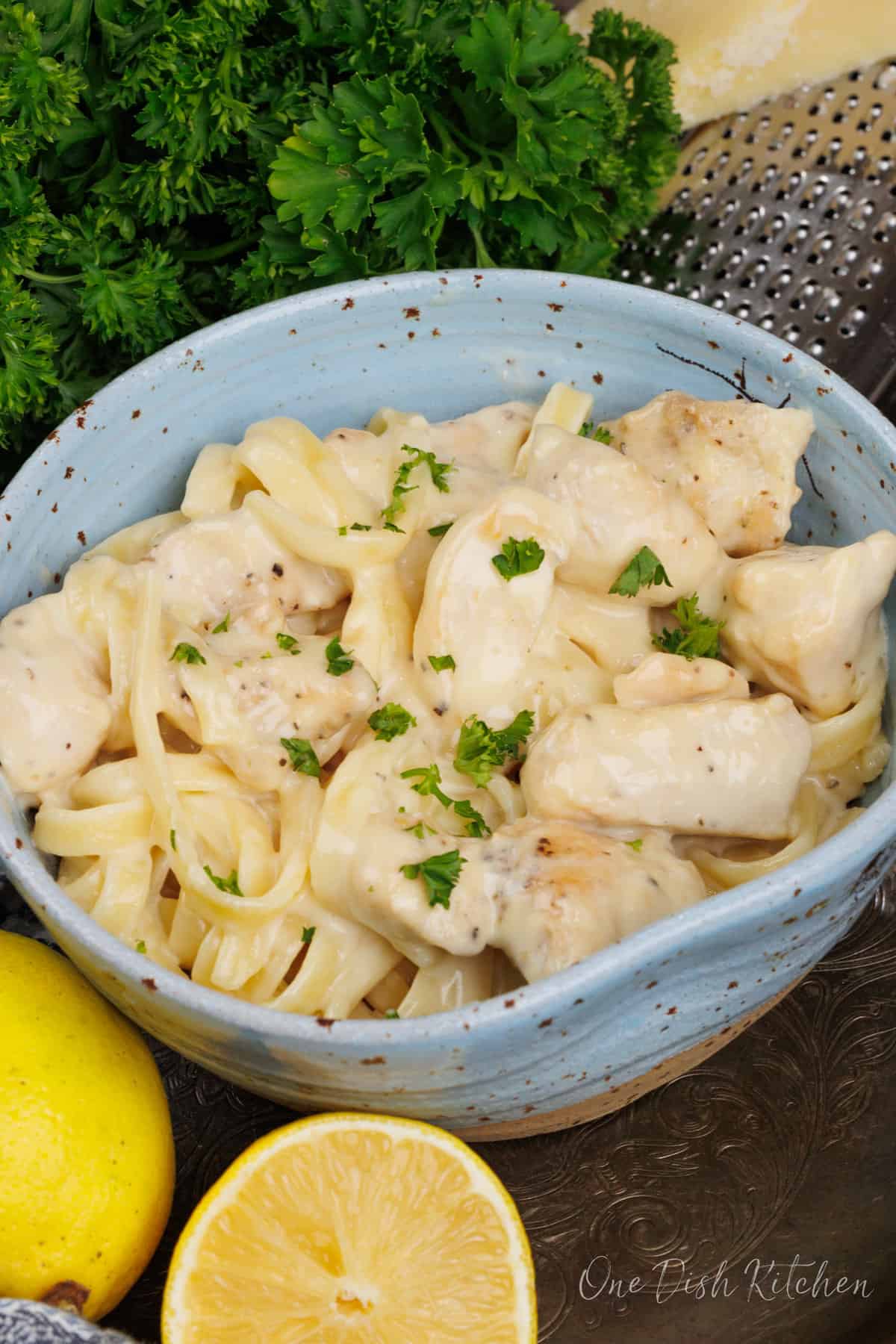 a blue bowl filled with chicken alfredo topped with chopped parsley and next to a fresh lemon and a wedge of Parmesan cheese.