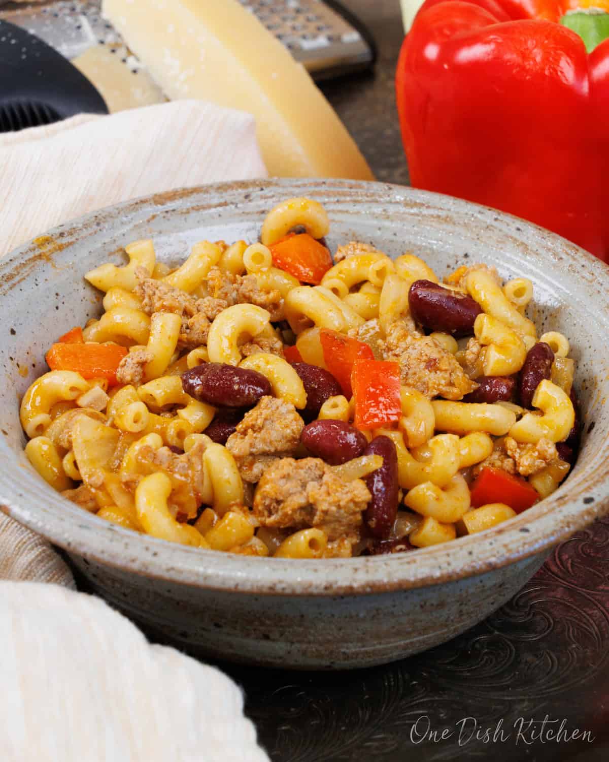 a blue bowl filled with chili mac on a silver tray next to a whole red bell pepper and a wedge of parmesan cheese.
