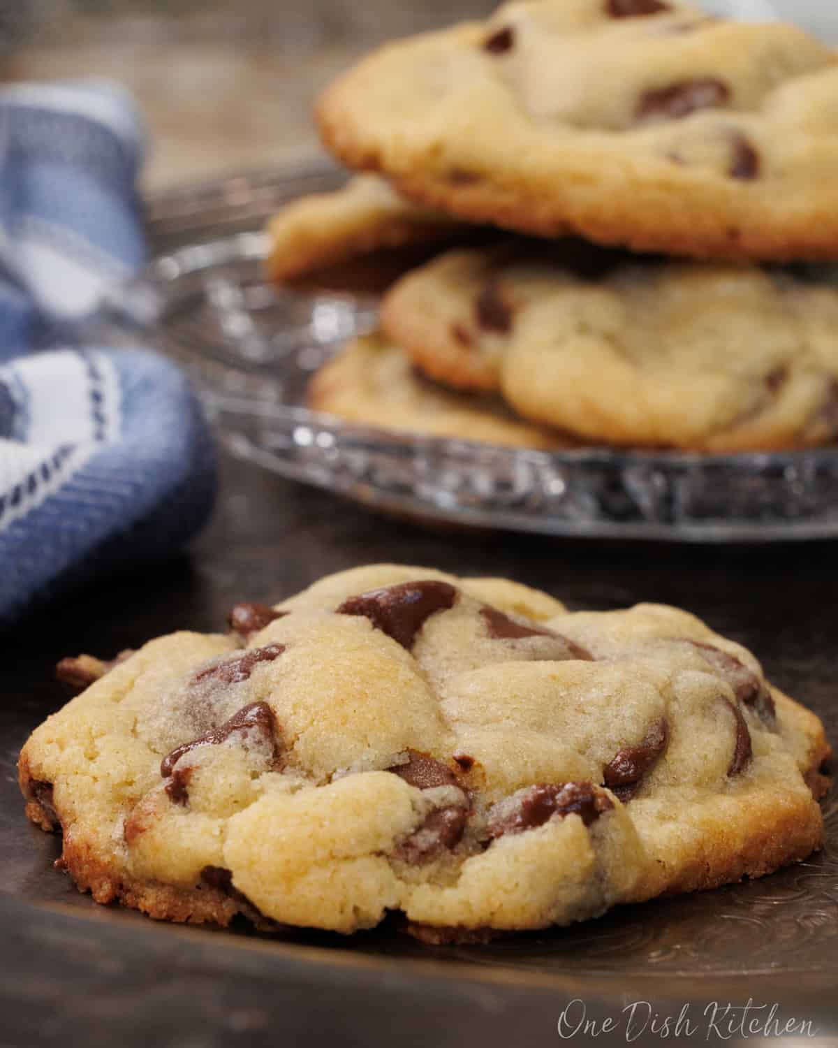 one chocolate chip cookie on a silver tray next to a plate of four chocolate chip cookies.