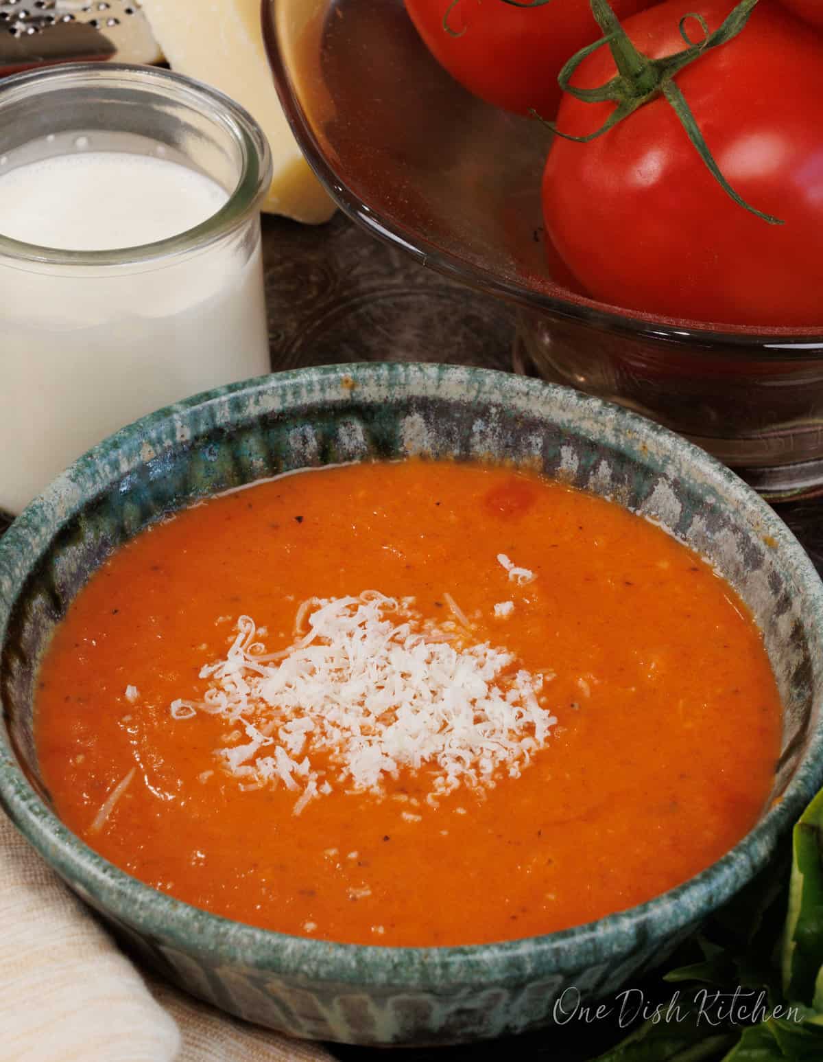 a green bowl filled with creamy tomato soup topped with grated parmesan cheese next to a bowl of fresh tomatoes