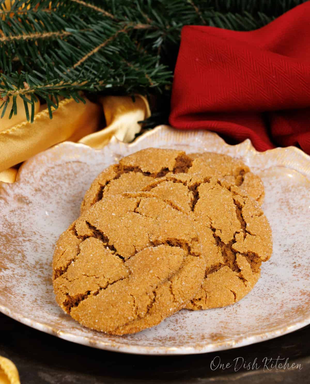 three molasses cookies on a white plate next to Christmas greenery and a red napkin on a silver tray