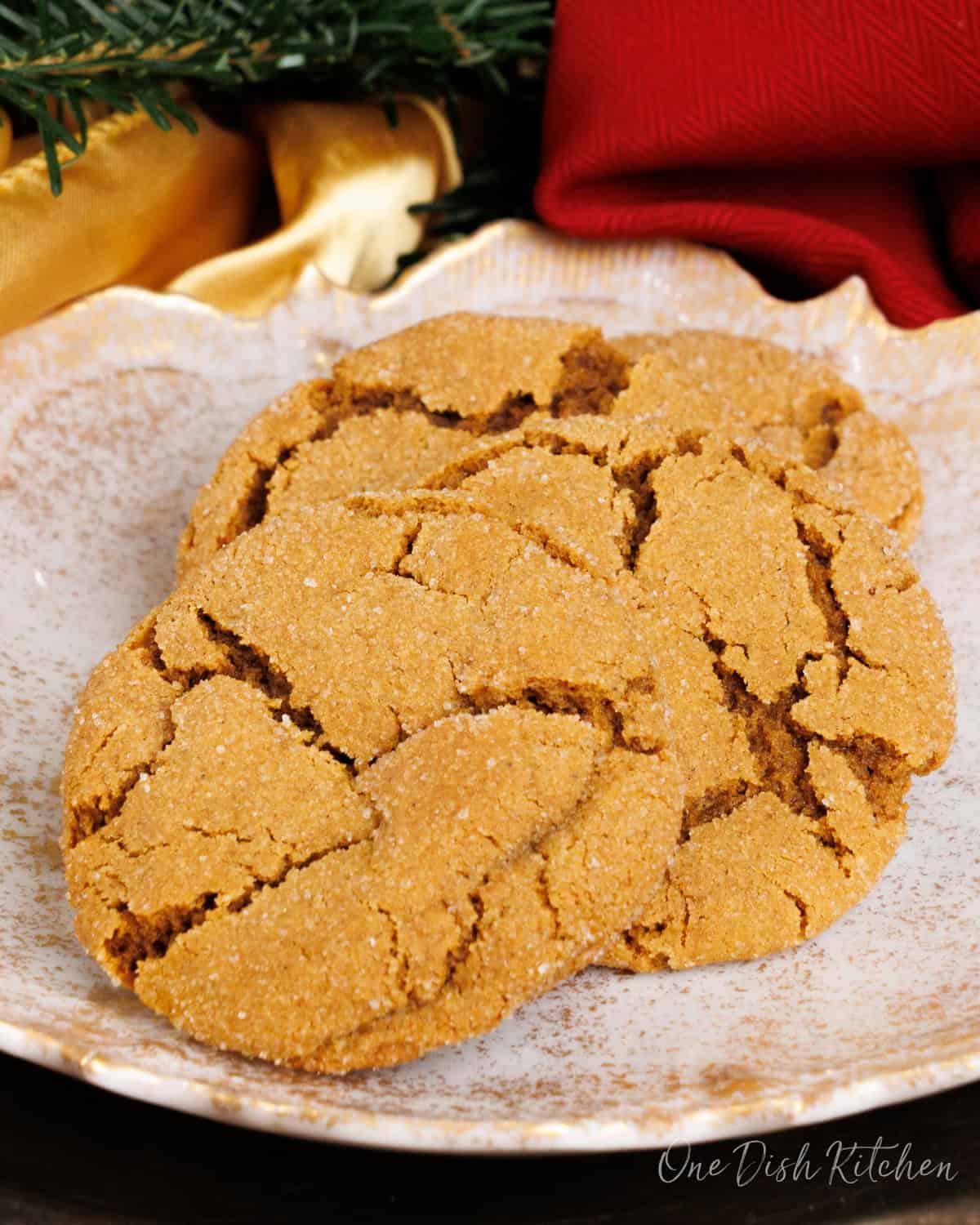 three molasses cookies on a white plate next to Christmas greenery and a red napkin on a silver tray.