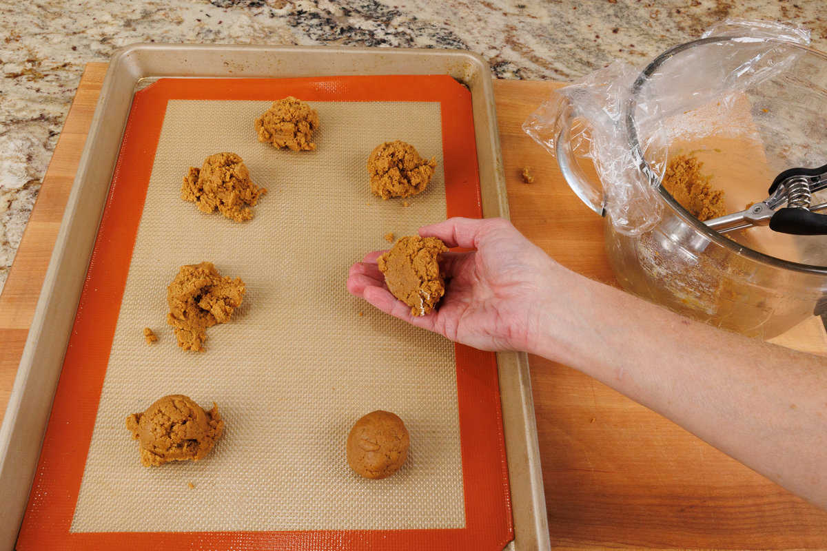 scooping molasses cookie dough from a mixing bowl and placing the balls on a lined baking sheet