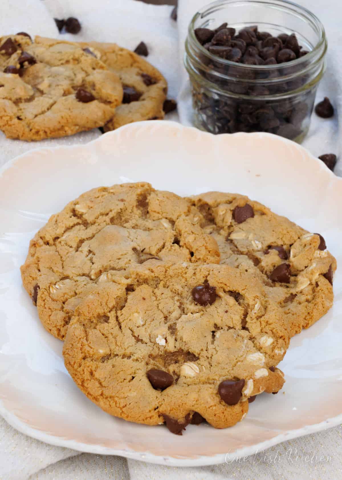 three oatmeal chocolate chip cookies on a plate next to a jar of chocolate chips and a white napkin