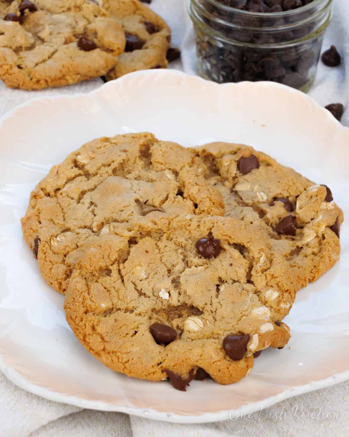 three oatmeal chocolate chip cookies on a plate next to a jar of chocolate chips and a white napkin.