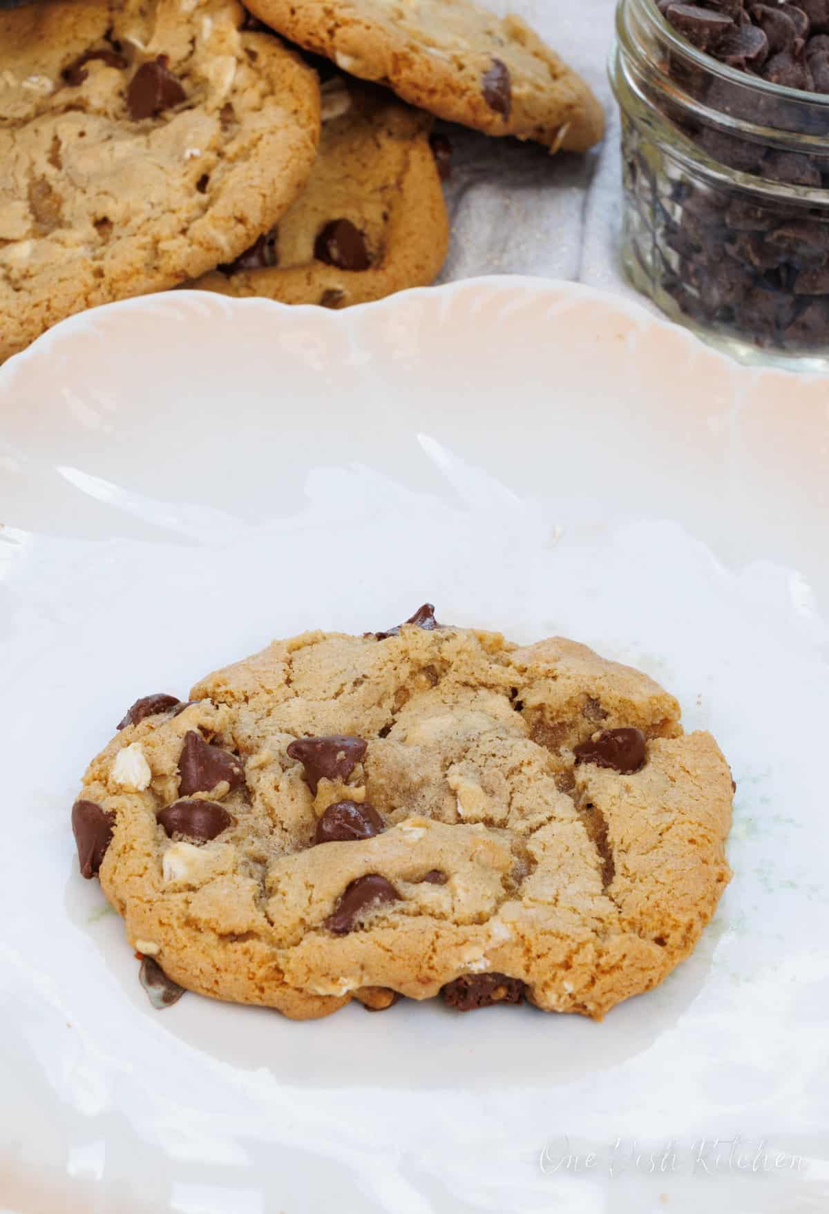 one oatmeal chocolate chip cookie on a white plate next to three cookies and a bowl of chocolate chips