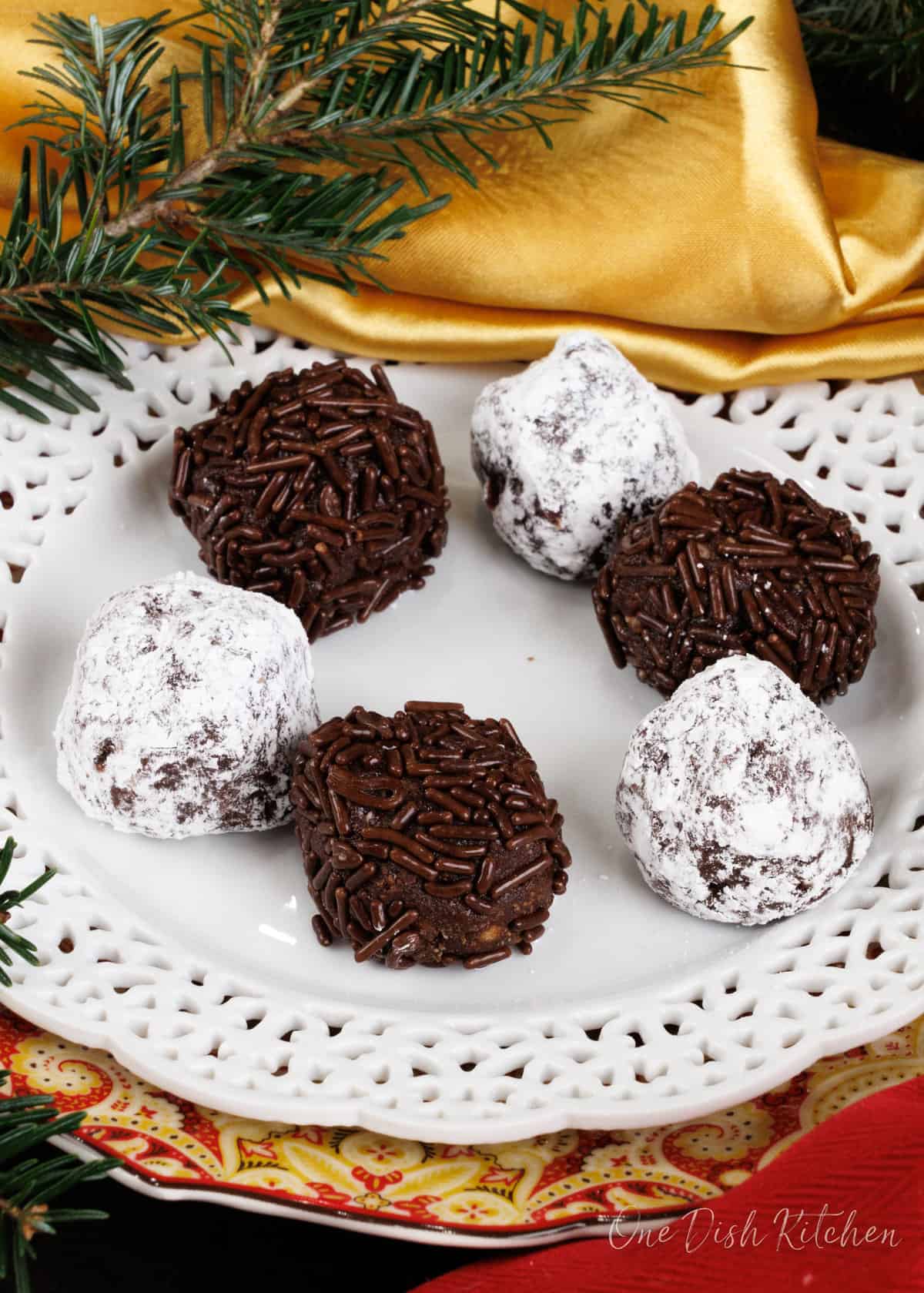 6 rum balls rolled in sprinkles and powdered sugar on a white plate next to a gold napkin and leaves from a tree.