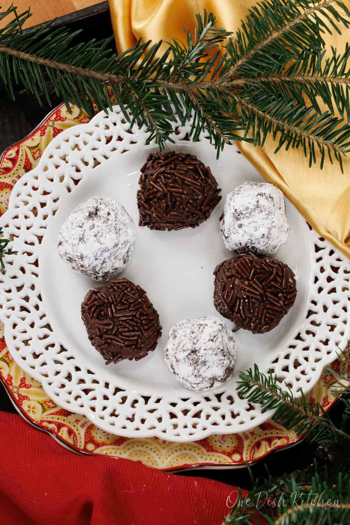 a overhead view of 3 rum balls rolled in powdered sugar and 3 rum balls rolled in sprinkles on a white plate next to a christmas tree sprig