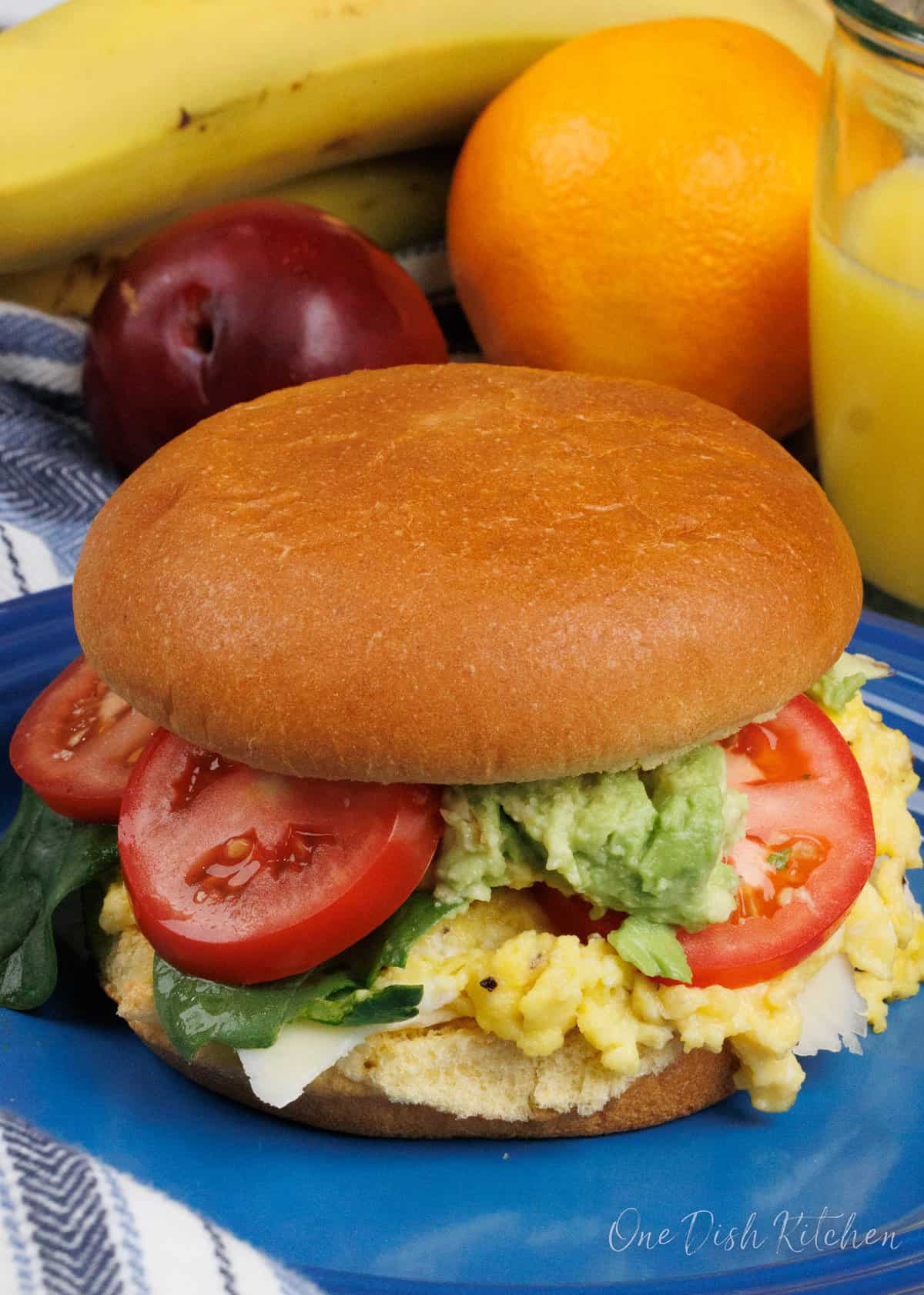 a bun filled with eggs, tomatoes, and an avocado on a blue plate next to a bowl of fruit and a glass of orange juice