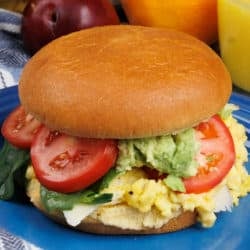 a bun filled with eggs, tomatoes, and an avocado on a blue plate next to a bowl of fruit and a glass of orange juice