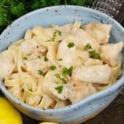 a blue bowl filled with chicken alfredo topped with chopped parsley and next to a fresh lemon and a wedge of Parmesan cheese.