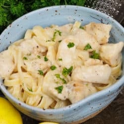 a blue bowl filled with chicken alfredo topped with chopped parsley and next to a fresh lemon and a wedge of Parmesan cheese.
