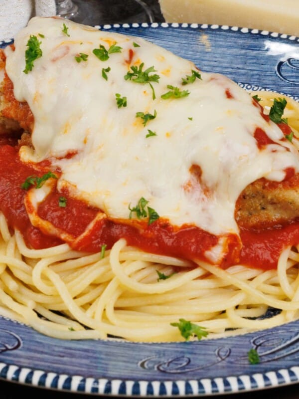a plate of chicken parmesan served over spaghetti next to a white napkin and a wedge of Parmesan cheese